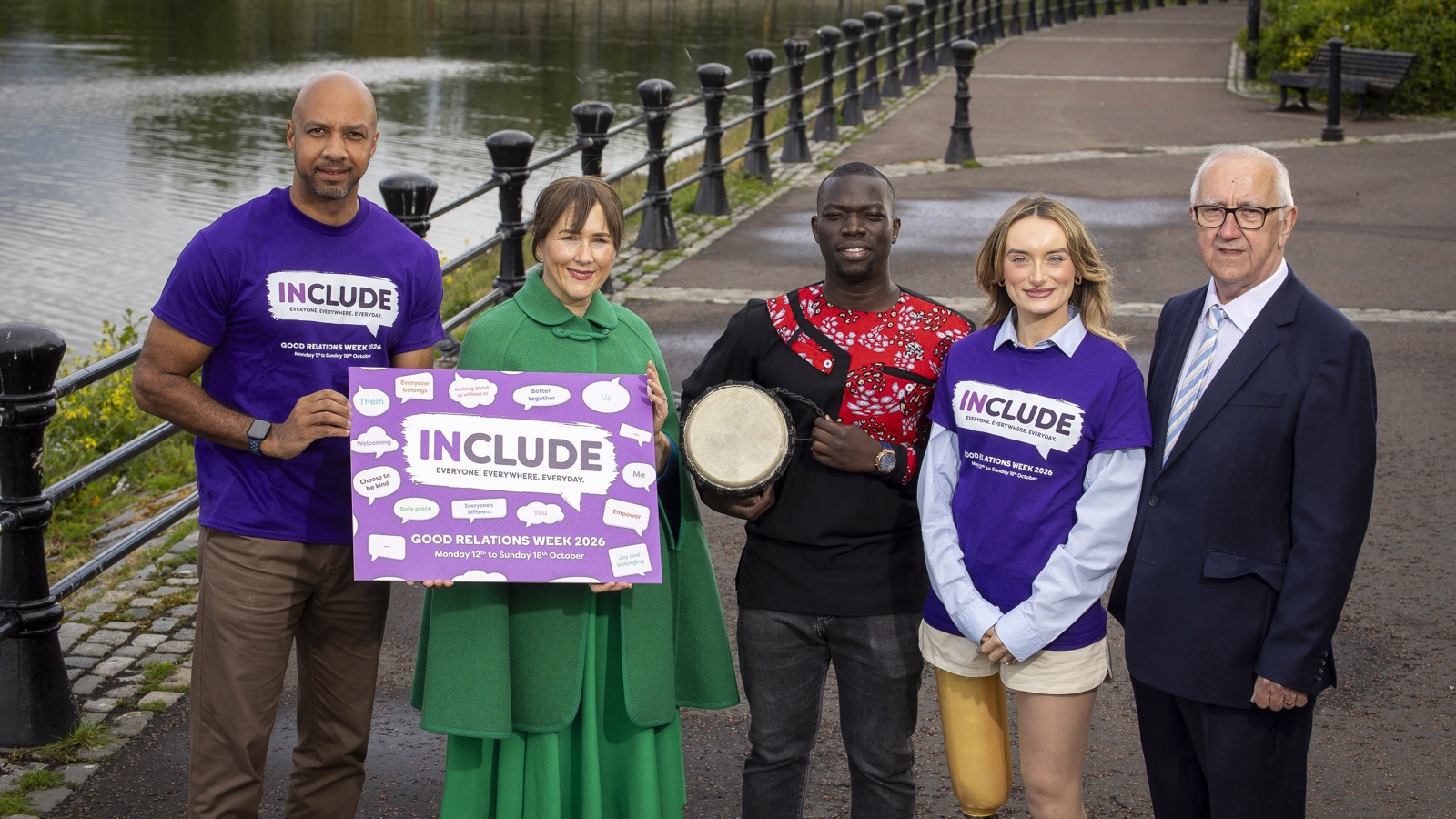 Five people on a riverside path at the Good Relations Week 2026 launch, three in purple Include T-shirts, one holding the Include poster and one a drum.